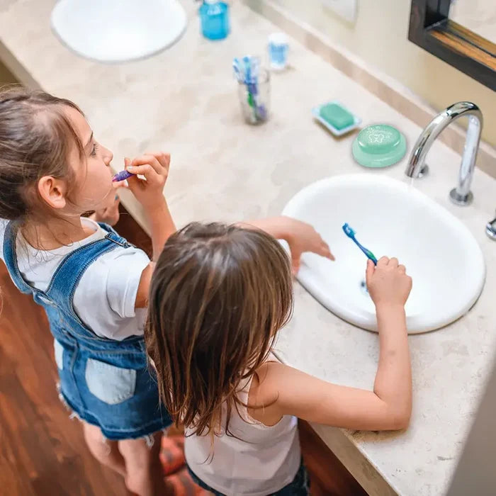 Round green light-up timer with a white tooth graphic on a bathroom counter as two young girls brush their teeth.