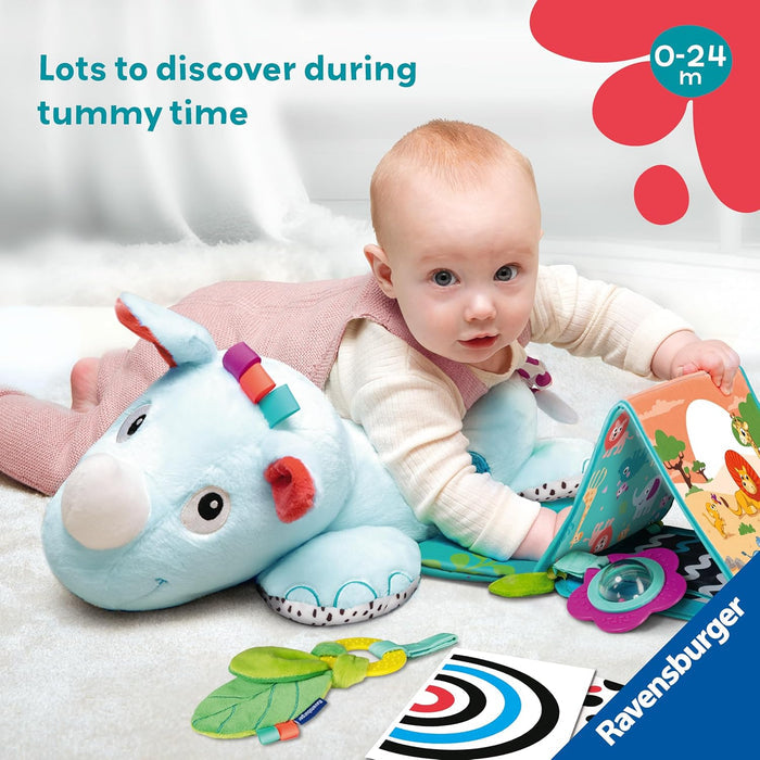 A baby on tummy time using a plush blue rhino pillow with a fabric book, rattles, and high-contrast sensory cards.