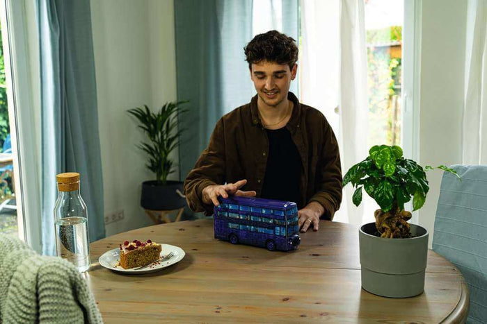 Purple triple-decker bus 3D puzzle on a wooden table, shown in a lifestyle setting with a person and cake.