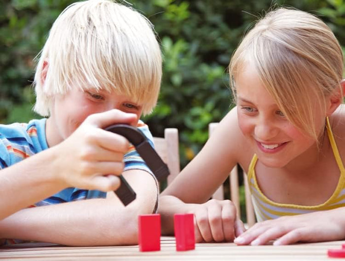Two children smiling as they use a large black horseshoe magnet on two small red rectangular blocks outdoors.