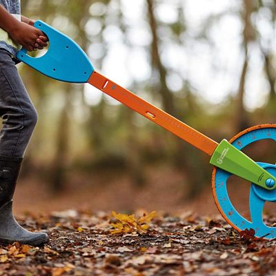 Child pushes a colorful trundle wheel with a blue handle and orange arm on a leaf-covered forest floor.