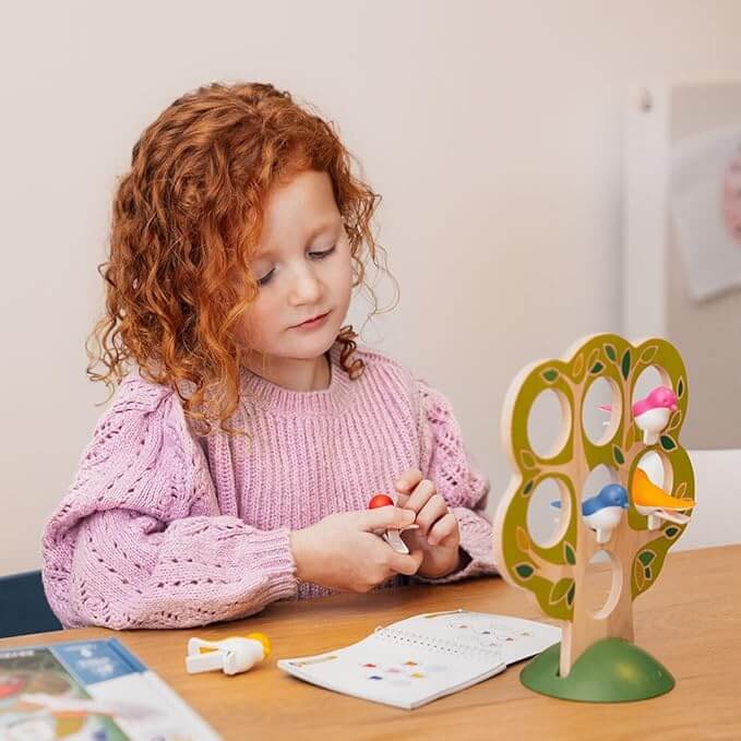 Red-haired girl attaches colorful bird clips to a wooden tree toy at a table with an open instruction booklet.