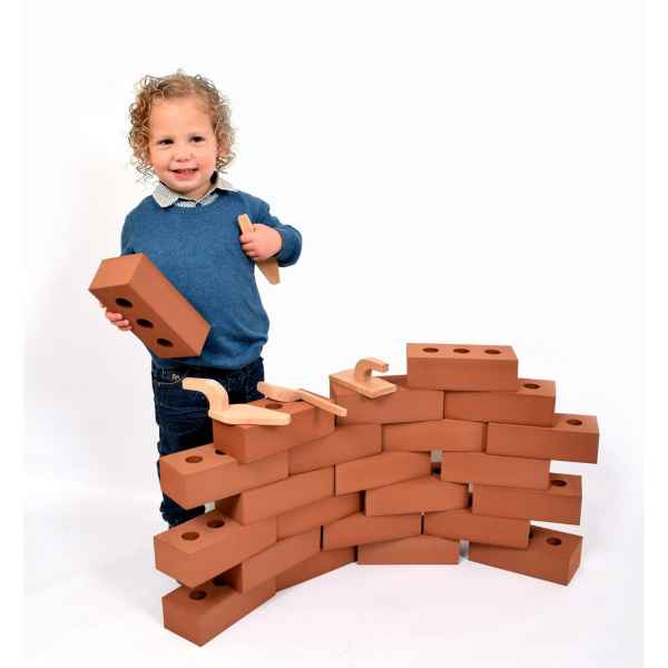 A young child building a curved wall using large, reddish-orange rectangular toy bricks and wooden play tools.