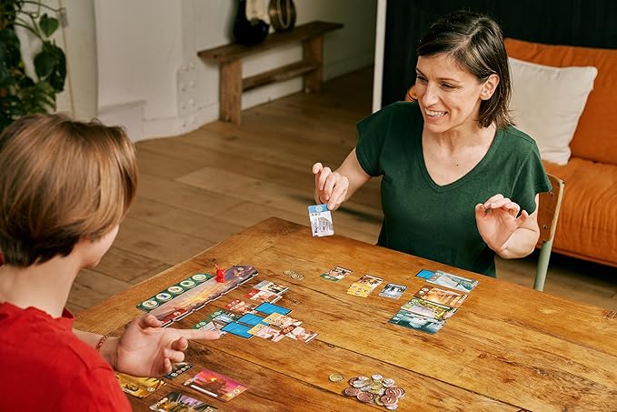 Two people playing a board game with colorful cards and silver tokens on a rustic wooden table.