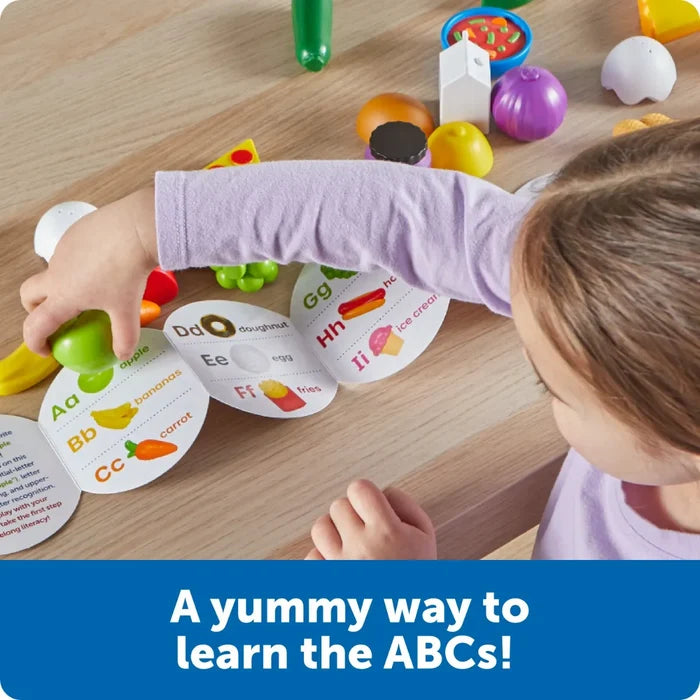 A child matches toy foods like a green apple and purple onion to an alphabet booklet on a wooden table.