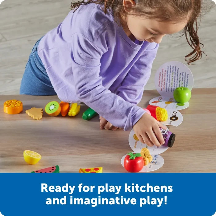 A child matches colorful toy foods like a kiwi and tomato to round alphabet letter cards on a wooden tabletop.