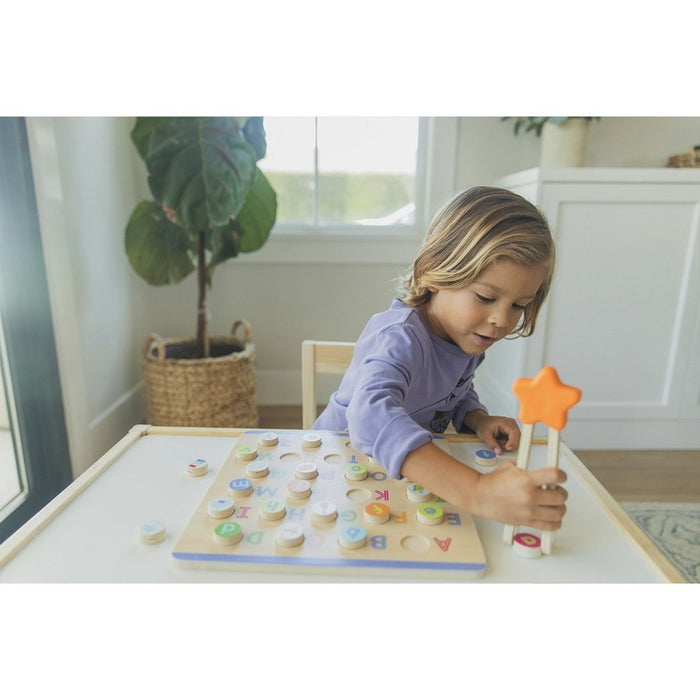 Child using a star-shaped wooden tool to pick up circular letter pieces on a colorful alphabet puzzle board.