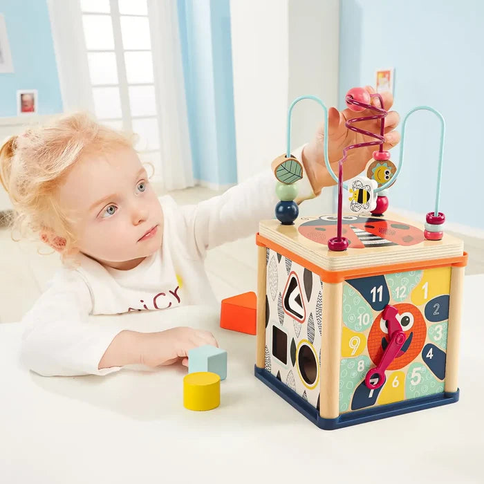 Young girl playing with a colorful wooden activity cube featuring a bead maze, clock face, and shape sorter.