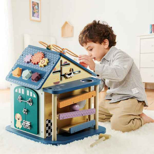 Child playing with a wooden activity house featuring colorful gears, shape sorters, and a ball ramp on a white rug.