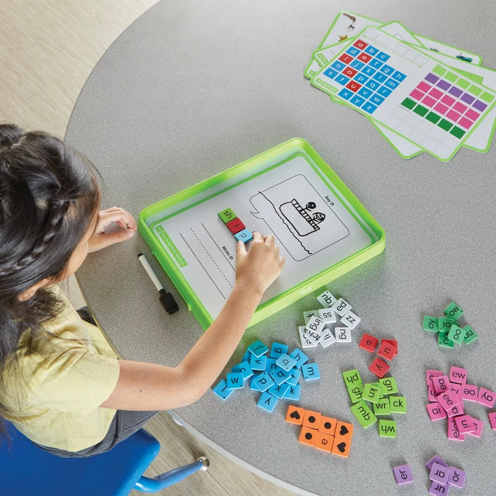 Child spells ship using colorful letter tiles on a green magnetic tray with various phonics tiles on a table.