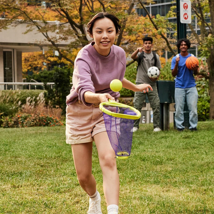 A young woman catching a yellow ball with a lime green and purple mesh handheld net toy in a grassy park.