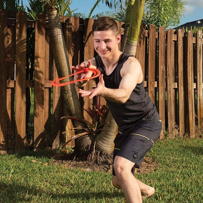 A man in a black tank top and shorts crouches outdoors in a backyard, reaching for a red plastic ring-shaped toy in his hands. A wooden fence, palm trees, and green grass are visible in the background.