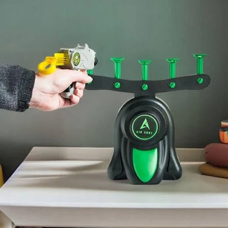 Hand aiming a gray toy gun at a black and neon green target base with four air nozzles on a white tabletop.