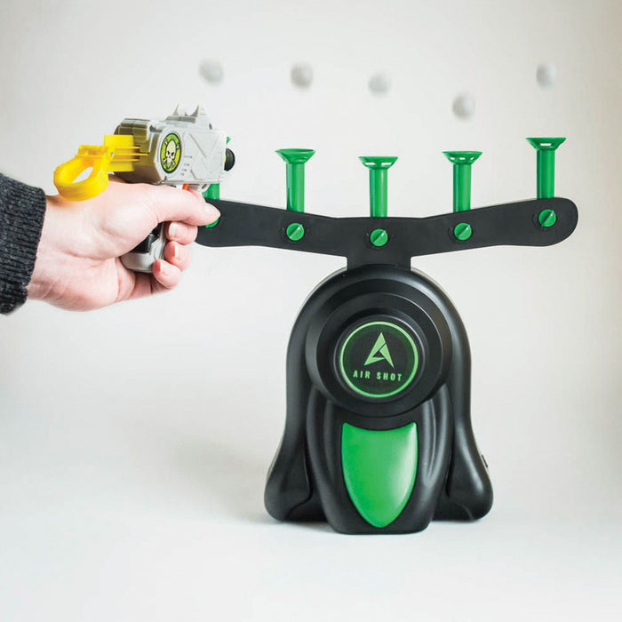 Hand holds a toy blaster aiming at a black and green target base with four white foam balls hovering in mid-air.