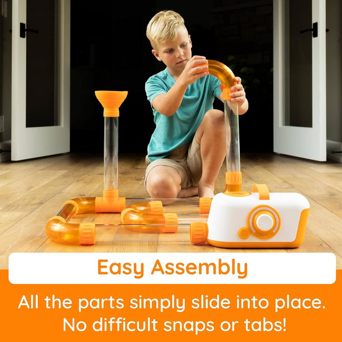 Child connects orange elbow joints to clear tubes with a white air-powered base on a wooden floor.