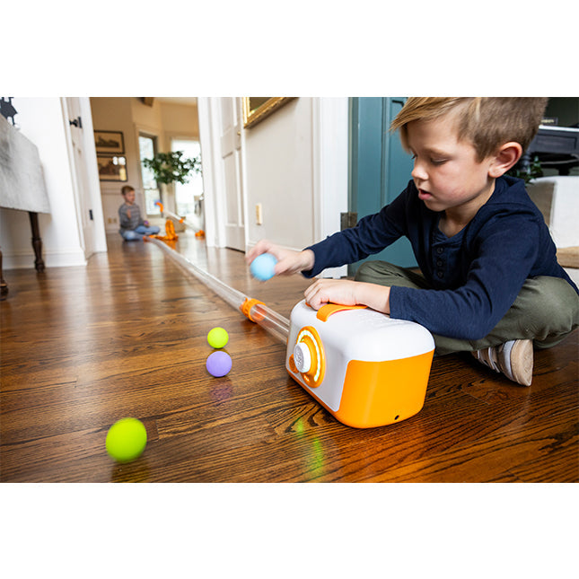A child plays with an orange air launcher and long clear tube extension, sending foam balls down a hallway.