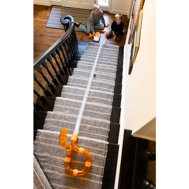 Long clear tube with orange curved ends extends down carpeted stairs as a blue ball travels through.