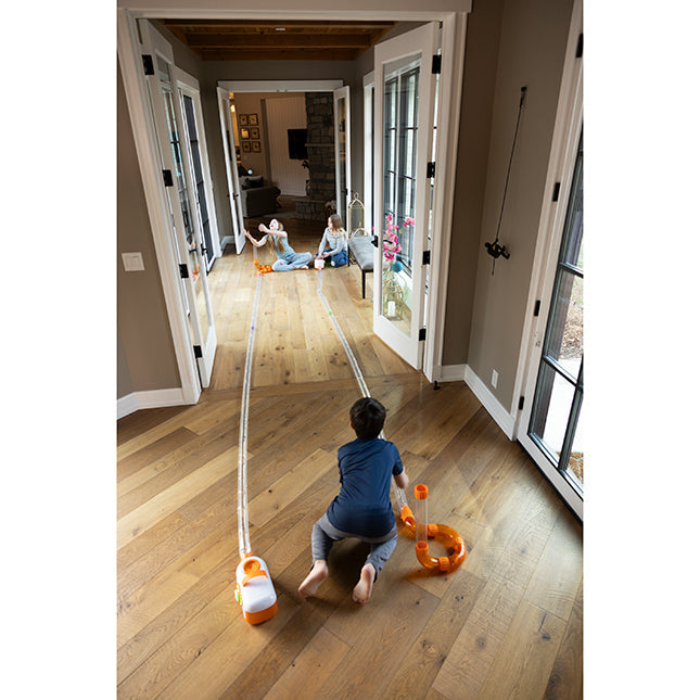 Three children play with long clear tubes and orange connectors stretching across a long hallway floor.