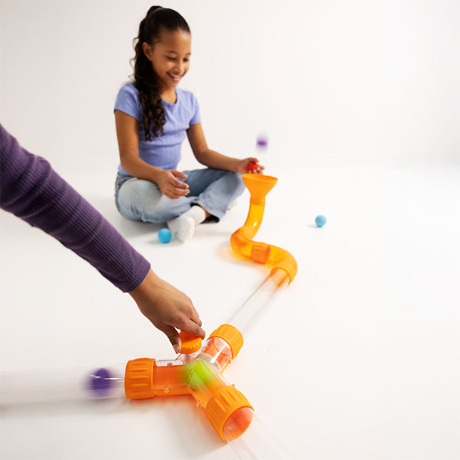 Hand turns an orange knob on a Y-shaped tube connector joined to clear tubes as a purple ball moves through.
