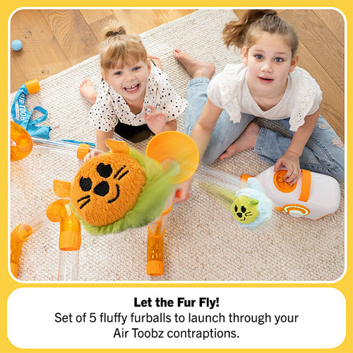 Two girls launching plush orange and blue furballs with faces through a clear tube system and white air blower.