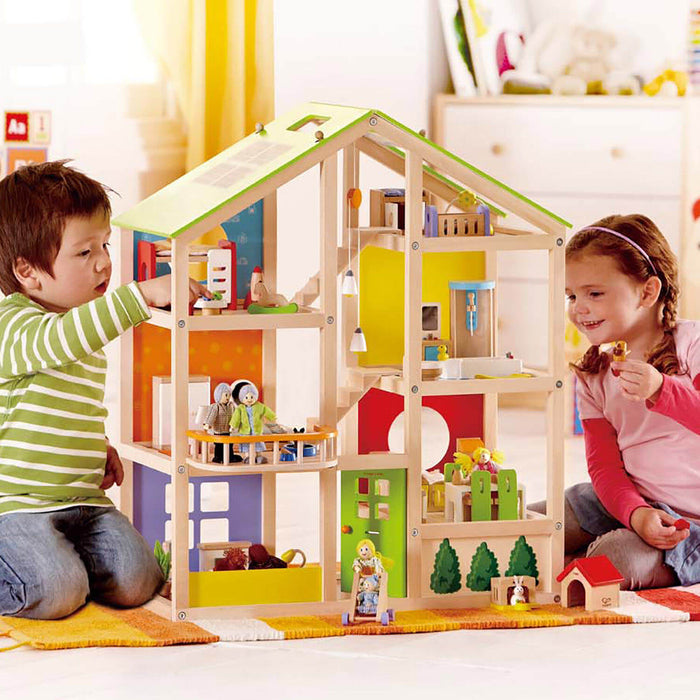 Two children playing with a three-story wooden dollhouse featuring a green roof, solar panels, and colorful furnishings.