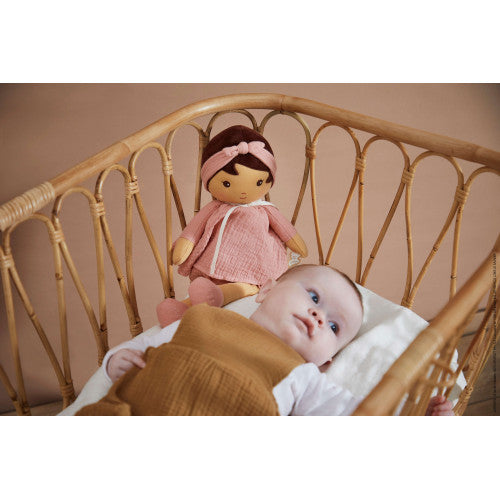 Soft plush doll with pink dress and headband in a rattan bassinet next to an infant wearing a brown outfit.