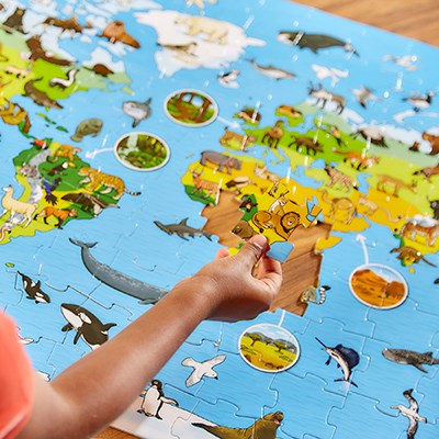 Hand placing a lion piece into a large blue world map puzzle featuring colorful illustrations of animals.