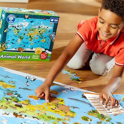 Smiling boy in orange shirt assembling a large animal world map jigsaw puzzle on a floor next to its green box.