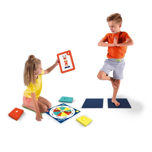 A girl shows an animal yoga card to a boy balancing on one leg on a foam mat, surrounded by a spinner and cards.