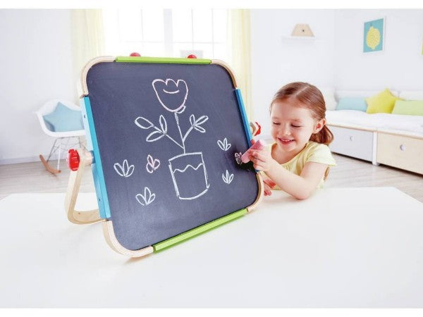 A young girl drawing a flower on a wooden tabletop chalkboard easel with pink chalk in a bright playroom.
