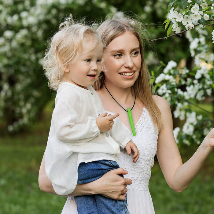 Woman wearing a lime green, saber-shaped sensory chew pendant on a black cord while holding a toddler in a garden.