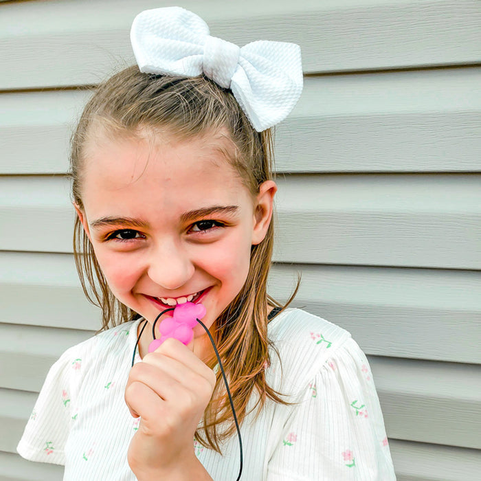 Smiling girl biting a pink gummy bear-shaped sensory chew necklace on a black cord.