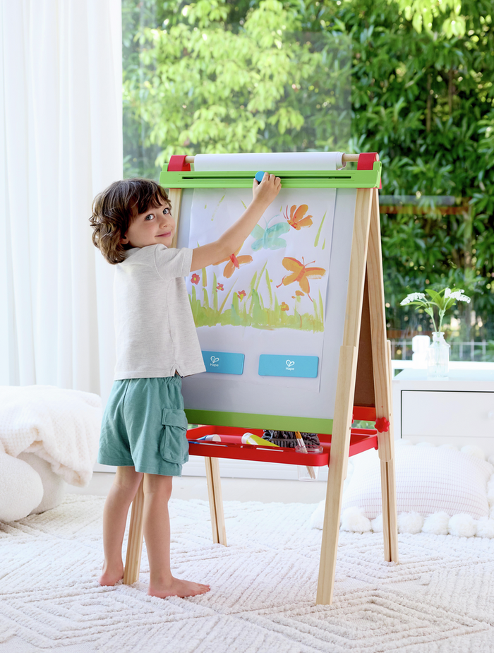 Child painting butterflies on a wooden easel with red and green trim, a paper roll, and a bottom supply tray.