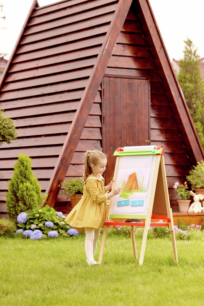 A child paints on a wooden A-frame easel with green trim and a paper roll in a garden with a wooden playhouse.