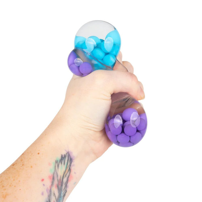 Hand squeezing a clear, flexible toy filled with small blue and purple beads, causing the ends to bulge.