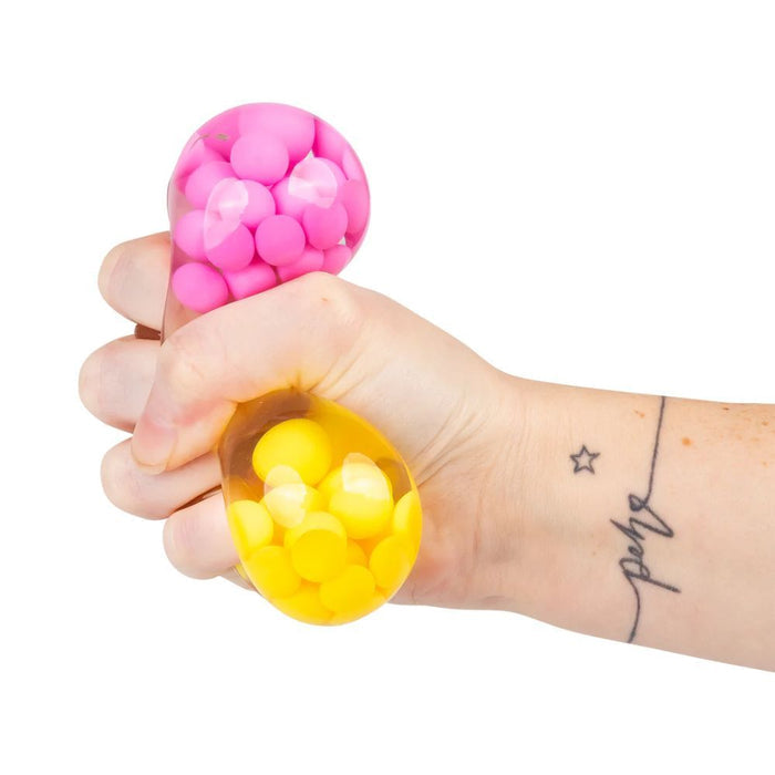 A hand squeezes a clear stress ball filled with small, bright pink and yellow spheres.