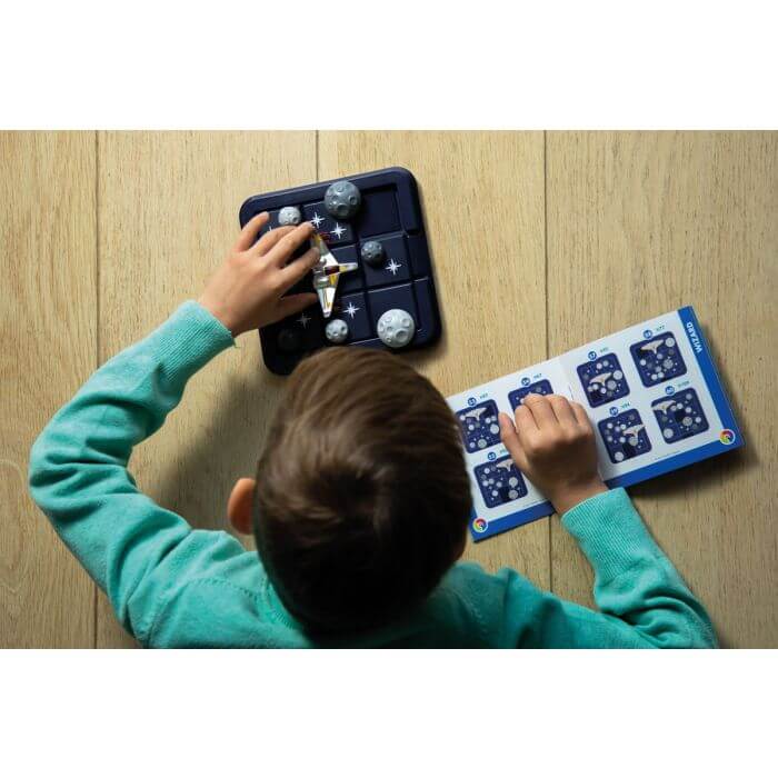 Child moves a spaceship on a blue grid board with grey asteroids, following an open instruction booklet.