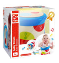 Toy drum in packaging showing a baby playing, with a blue rim, wooden top, and colorful musical note patterns.