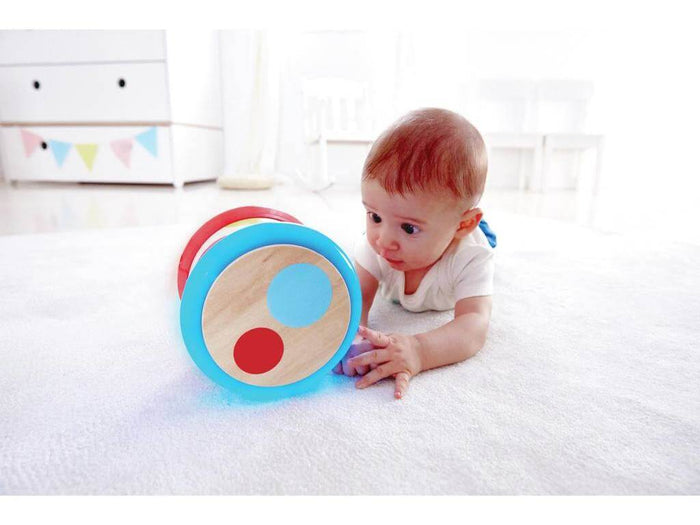 Baby on a rug reaches for a round wooden drum with a light blue rim and red and blue circles on its face.