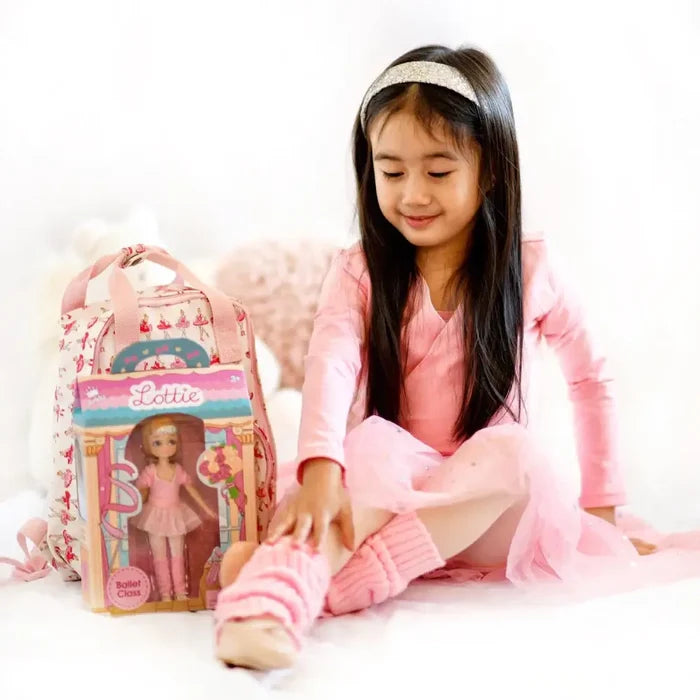 Girl in a pink ballet outfit sits next to a boxed doll and a pink ballerina-patterned backpack on a white background.