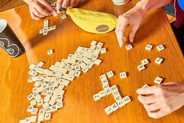 Yellow banana-shaped pouch and white letter tiles in crossword patterns on a wooden table as players arrange pieces.