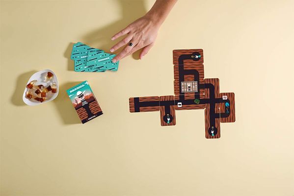 Hand reaching for green cards next to interlocking tunnel path tiles and a bowl of snacks on a tan surface.