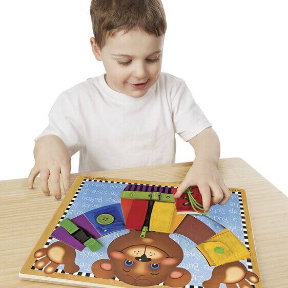 Boy interacts with a wooden teddy bear puzzle featuring colorful clothing with a zipper, buckle, laces, and snap.