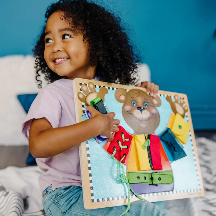 Smiling girl holds a wooden bear activity board with colorful clothes, a zipper, belt buckle, and laces.