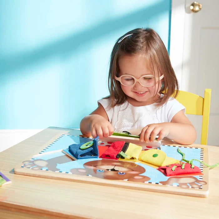 A girl playing with a wooden activity board featuring colorful fabric pieces for zipping, buttoning, and lacing.
