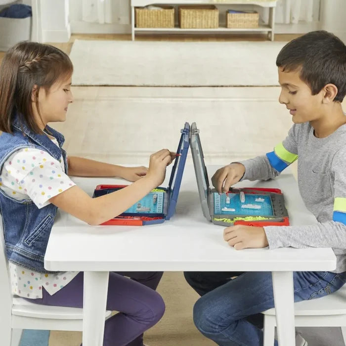 Two children playing Battleship at a white table with folding red and blue plastic cases, grid boards, and mini ships.