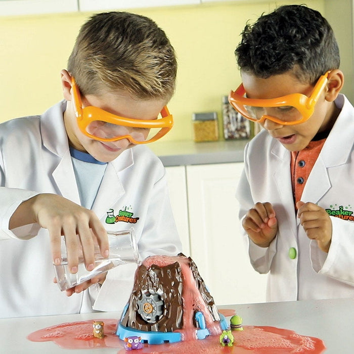 Boys in lab coats and orange goggles pour water into a brown toy volcano erupting pink foam and small figurines.