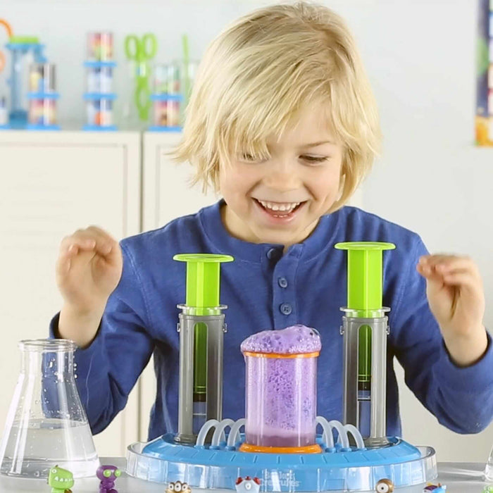 Smiling boy creates bubbling purple foam with a blue science toy featuring lime green plungers and tiny monsters.