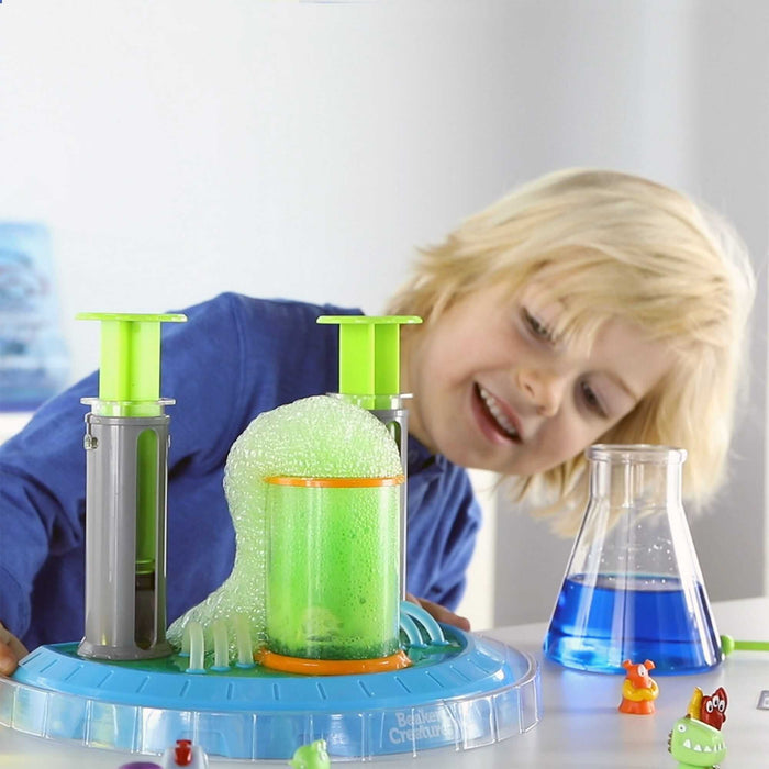 Blond child watches green foam bubble from a science lab toy with a blue base and blue liquid in a glass beaker.