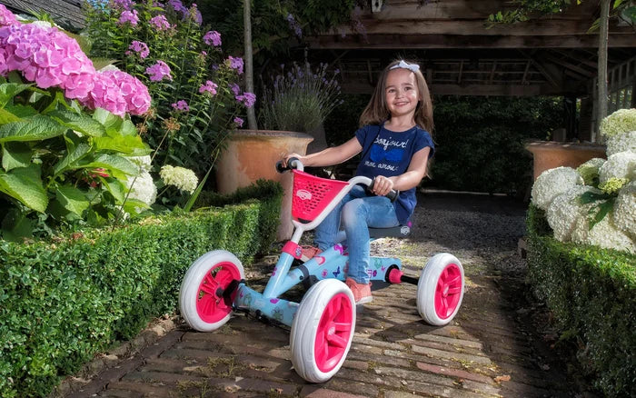 Light blue 4-wheel pedal go-kart with pink floral decals and basket, ridden by a girl on a brick garden path.